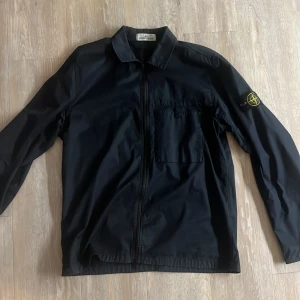 Svart jacka från Stone Island - Snygg svart jacka från Stone Island med dragkedja och långärmad design. Jackan har en broderad logga på ärmen och en klassisk krage. Perfekt för en stilren look. Nypris 2500-4000kr. 