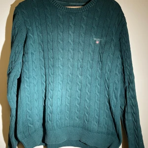 Grön kabelstickad tröja från Gant - Säljer en stilren grön kabelstickad tröja från Gant i premium bomull. Tröjan har en klassisk rund hals och långa ärmar, perfekt för kyligare dagar. Den har en diskret logga på bröstet och ribbade muddar för en bekväm passform.