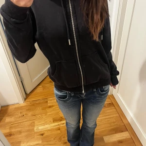Svart hoodie med dragkedja - Säljer en svart hoodie med dragkedja och fickor framtill. Den har en justerbar huva med snören och långa ärmar. 