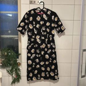 Blommig klänning från Camilla Thulin - Säljer en svart klänning från Camilla Thulin med vackert blommönster i vitt och gult. Klänningen har knappar framtill och trekvartsärmar, vilket ger en elegant look. Perfekt för vår och sommar!