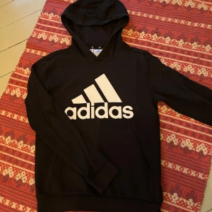 Svart hoodie från Adidas - Säljer en snygg svart hoodie från Adidas med den klassiska loggan i vitt på framsidan i bra skick.