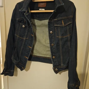 Mörkblå jeansjacka från Gul & Blå - Snygg mörkblå jeansjacka från Gul & Blå med klassisk design. Jackan har knappar framtill och två bröstfickor med knappstängning. Perfekt för en avslappnad stil.