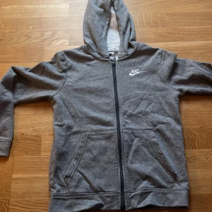 Grå hoodie från Nike - Säljer en grå hoodie från Nike med dragkedja och broderad logga på bröstet. Tröjan har en bekväm passform med långa ärmar och praktiska fickor framtill. Perfekt för en avslappnad stil.