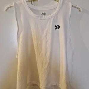 Vit ärmlös topp från Lager 157 - Säljer en vit ärmlös linnetopp med rund halsringning och en liten svart logotyp på bröstet. Perfekt för träning eller varma sommardagar.
