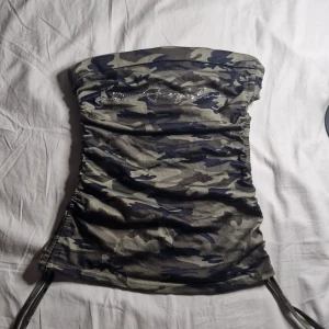 Camouflage tubtopp med rhinestones - Snygg camouflage tubtopp med glittrande strassdetaljer framtill. Perfekt för en cool och avslappnad stil. Tubtoppen har en åtsittande passform och är tillverkad i ett mjukt material.