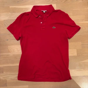 Röd pikétröja från Lacoste - Kolla sista bild för storlek.  Snygg röd pikétröja från Lacoste med klassisk krage och knappar framtill. Tröjan har det ikoniska krokodilmärket broderat på bröstet. Perfekt för en stilren och avslappnad look.