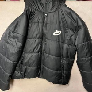 Svart pufferjacka från Nike - Snygg svart pufferjacka (medium) för ungdom 10-13 år. Märke Nike med dragkedja och huva. Jackan har ett klassiskt quiltat mönster och Nike-logga på bröstet och ryggen. Perfekt för kyliga dagar på fotbollsplanen tex.