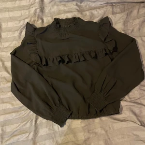 Svart blus med volangdetaljer - Snygg svart blus med volangdetaljer på bröstet och axlarna. Den har långa ärmar med elastiska muddar och en hög krage med smock. Perfekt för en stilren look.