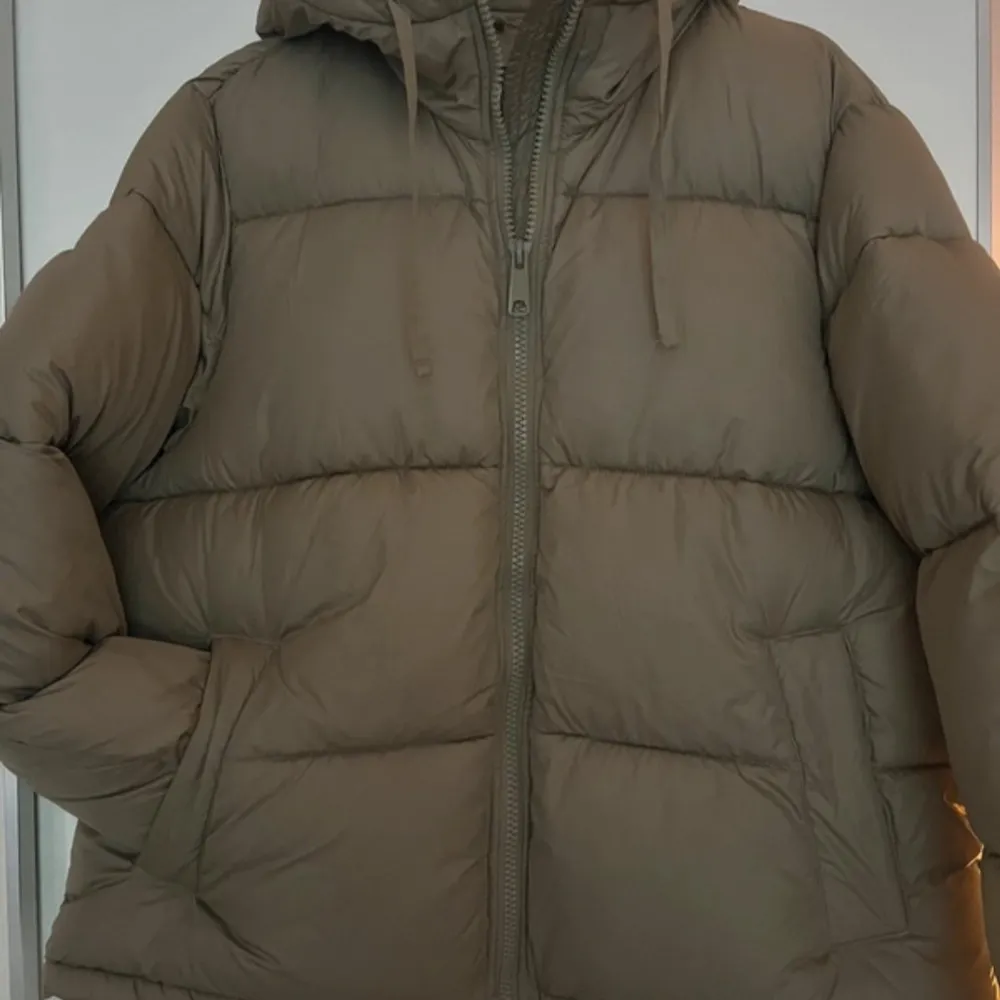 Säljer en mysig beige/grön dunjacka från H&M. Jackan har en dragkedja framtill och en stor huva för extra värme. Perfekt för kyliga dagar med sin pufferdesign som ger en skön och avslappnad look.. Takit.