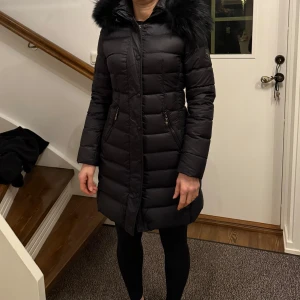 Svart dunjacka med pälskrage - Snygg svart vinterjacka från Hollies med en pälskrage. Jackan har en figursydd passform och stängs med dragkedja, luvan är även avtagbar med dragkedja. 