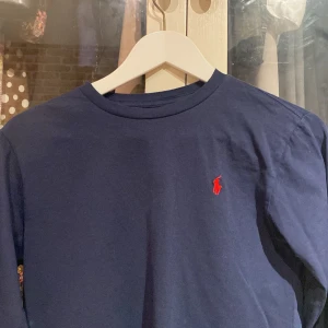 Mörkblå långärmad tröja från Polo Ralph Lauren - Säljer en stilren mörkblå långärmad tröja från Polo Ralph Lauren med det klassiska röda logotypen på bröstet. Perfekt för en avslappnad och snygg look. Tröjan är tillverkad i mjukt bomullsmaterial.