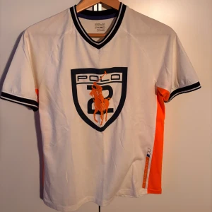 Vit kortärmad t-shirt från Polo Ralph Lauren - Snygg vit t-shirt från Polo Ralph Lauren med korta ärmar och kontrasterande orange detaljer på sidorna. Den har en stor broderad logga på framsidan. Storlek L i barn men passar perfekt för S. Använd 3-4 gånger.