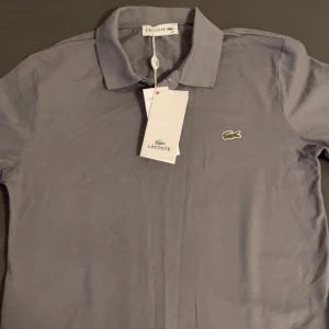 Grön pikétröja från Lacoste - Säljer en stilren grå pikétröja från Lacoste med den klassiska krokodilloggan på bröstet. Tröjan har en krage och korta ärmar, perfekt för en avslappnad men ändå snygg look. Passar bra till både jeans och chinos. 🐊