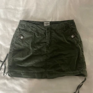 Grön kortkjol med knappar - Snygg grön kortkjol med praktiska fickor och knappar både fram och bak. Kjolen har en avslappnad stil med snörning i sidorna för justerbar passform. Storlek 40/L