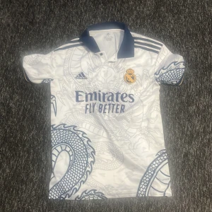 Vit och blå Real Madrid fotbollströja - Snygg vit och blå Real Madrid fotbollströja från Adidas med drakmönster. Tröjan har korta ärmar och klubbens emblem på bröstet. Perfekt för fans som vill visa sitt stöd med stil.