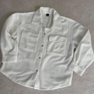 Vit jacka från Ginatricot - Säljer en stilren vit overshirt från Ginatricot. Den har en klassisk krage och knappar framtill. Två stora fickor på bröstet ger en cool look. Perfekt för lager-på-lager-stil under hösten eller våren.