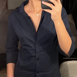 Mörkblå skjorta med knappar - Säljer en stilren mörkblå skjorta med knappar. Skjortan har trekvartsärmar och en klassisk krage. Perfekt för både vardag och festliga tillfällen. Passar bra till jeans eller kjol för en snygg look! Skjortan är i nyskick 