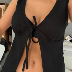 Svart ribbad topp med knytning - Säljer en snygg svart ribbad topp som är ärmlös och har en cool knytning framtill. Perfekt för sommaren eller en utekväll. Toppen har små volanger längst ner på tröjan och ger en trendig look.