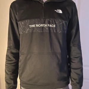 Svart tröja från The North Face - Säljer en svart tröja från The North Face med dragkedja vid halsen. Tröjan har ett tryck med The North Face-loggan över bröstet och är perfekt för både vardag och utomhusaktiviteter. Den är långärmad och har en normal passform.