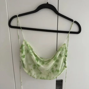 Grön blommig linnetopp - Säljer en superfin grön blommig linnetopp från NAKD, Hanna Schönbergs kollektion. Den har tunna axelband och ett löst, draperat fall framtill. Helt öppen i ryggen, endast knyte. Perfekt för sommardagar eller en avslappnad festkväll. Toppen är i nyskick 