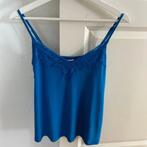 Blå ärmlös topp med spetsdetaljer - Säljer en snygg blå ärmlös topp med spetsdetaljer vid halsringningen. Perfekt för sommardagar eller som ett lager under en kavaj. Toppen har smala axelband och är gjord i ett mjukt material som känns skönt mot huden.