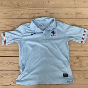 Ljusblå fotbollströja från Nike - Säljer en ljusblå fotbollströja från Nike med Frankrike loggan på bröstet, den är från säsongen 2013-2014 bortatröja . Tröjan är i Dri-FIT-material för att hålla dig sval och torr. Den har en klassisk krage och korta ärmar med ränder i rött och blått. Perfekt för fotbollsfans som vill ha en autentisk look! Pris går att diskutera