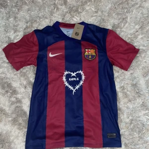 FC Barcelona fotbollströja - Säljer en FC Barcelona fotbollströja från Nike. Den är randig i rött och blått med klubbens emblem på bröstet och ett hjärtmönster med texten 'KARLG' på framsidan. Baksidan har UNHCR-loggan. Tröjan är kortärmad och i nyskick, perfekt för alla Barca-fans!