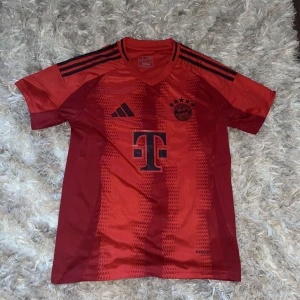 Röd FC Bayern München fotbollströja - Säljer en snygg röd FC Bayern München fotbollströja från Adidas. Tröjan har korta ärmar och är i nyskick, fortfarande i sin originalförpackning. Den har svarta detaljer och klubbens emblem på framsidan, samt texten 'FC Bayern München' på baksidan. Perfekt för alla Bayern-fans!