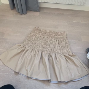 Beige kjol från Gina Tricot - Säljer en snygg beige kjol från Gina Tricot i storlek S. Kjolen har en elastisk midja med smockdetaljer och en volangkant som ger en fin form. Perfekt för att skapa en stilren look.