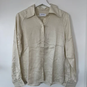 Beige skjorta från Rodebjer - Snygg beige skjorta från Rodebjer i en mjuk och glansig mix av cupro och rayon. Skjortan har långa ärmar och knappar framtill, perfekt för en stilren look. Den har en klassisk krage och en elegant passform.