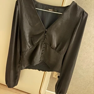 Svart blus från Bik Bok - Snygg svart blus från Bik Bok med v-ringning och knappar framtill. Blusen har långa ärmar och en elegant design som passar perfekt för olika tillfällen.