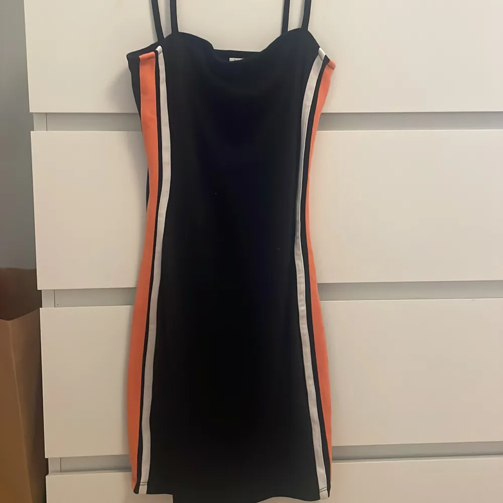Snygg svart klänning med smala axelband och kontrasterande orange och vita ränder längs sidorna. Perfekt för en cool och sportig look. Klänningen har en tight passform som framhäver figuren.. Mekot.