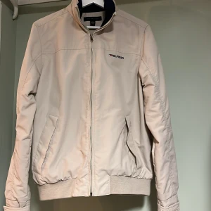 Beige jacka från Tommy Hilfiger - Snygg beige jacka från Tommy Hilfiger med dragkedja och ribbade muddar. Jackan har en diskret logga på bröstet och ärmarna. Perfekt för en stilren look. Limited Edition!