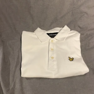 Vit pikétröja från Lyle & Scott - Säljer en stilren vit pikétröja från Lyle & Scott med korta ärmar och klassisk krage. Tröjan har en broderad logotyp i gult och svart på bröstet. Perfekt för en avslappnad och snygg look. Ej strykt på fotonen.