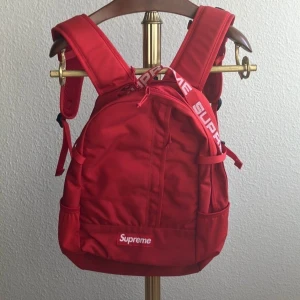 Röd ryggsäck från Supreme - Snygg röd ryggsäck från Supreme med justerbara axelremmar och flera praktiska fack. Väskan har Supreme-loggan på framsidan och axelremmarna, vilket ger en cool look. Perfekt för att bära dina grejer med stil.