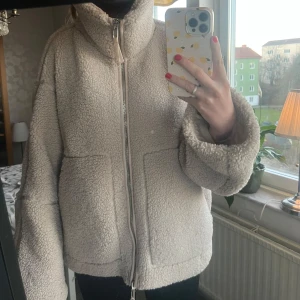 Beige teddyjacka med dragkedja - Mysig beige OVERSIZED teddyjacka från H&M med dragkedja och hög krage. Perfekt för kyliga dagar med sin fluffiga och varma design. Jackan har en avslappnad passform och stora fickor framtill. Jackan går att ha knäpp som en krage eller utan. 