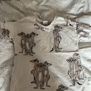 Beige t-shirt med surikatmönster från Lindex - Säljer en söt beige t-shirt från Lindex med ett lekfullt surikatmönster. Perfekt för små äventyrare! T-shirten har korta ärmar och är tillverkad i mjukt material för bekväm användning.