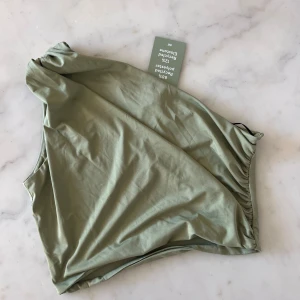 One shoulder topp - M helt NY från H&M - Säljer en stilren och trendig enaxlad topp från H&M i en fin olivgrön färg. Den har rynkade detaljer på sidorna och är gjord av ett mjukt, stretchigt material (88 % återvunnen polyester, 12 % elastan). Perfekt för både vardag och fest!