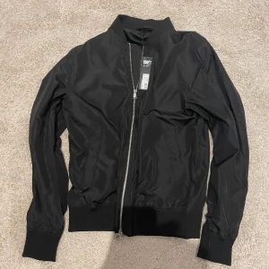 Svart bomberjacka från J.Lindeberg - Snygg svart bomberjacka från J.Lindeberg i storlek M. Jackan har en stilren design med dragkedja framtill och fin designade rubbar vid ärmslut och nederkant. Perfekt för en cool och avslappnad look. Den osar dig som är 170-178 cm och kan pass dig som är 180 cm lång. Jackan kommer direkt från butik helt orörd och oanvänd 