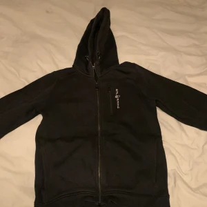 Svart hoodie från Sail Racing - Säljer en svart hoodie från Sail Racing med dragkedja framtill. Den har en klassisk design med huva och långa ärmar. Perfekt för kyliga dagar.