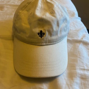 Beige Morris keps - Snygg beige keps med en broderad fleur-de-lis detalj framtill. Perfekt för en avslappnad stil och skydd mot solen. Justerbar passform för bekvämlighet.