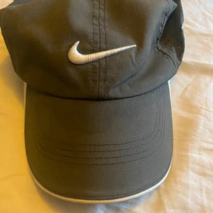 Grå keps från Nike - Snygg grå keps från Nike med vit broderad logga framtill. Kepsen har perforerade sidor för ventilation och texten '20XI' på sidan. Perfekt för sportiga aktiviteter.