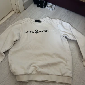 Vit tröja från Sail Racing - Säljer en vit tröja från Sail Racing med svart logotyp på bröstet. Tröjan har långa ärmar och en rund halsringning. Perfekt för en avslappnad stil.
