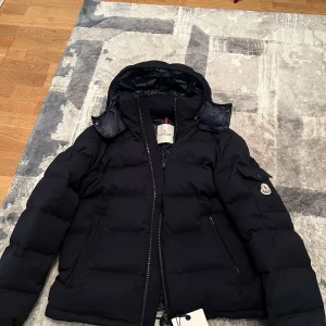 Svart dunjacka från Moncler - Säljer en snygg svart dunjacka från Moncler. Jackan har en dragkedja framtill och en avtagbar huva. Insidan är quiltad med ett coolt serietidningsmönster. Perfekt för kalla dagar! 🖤