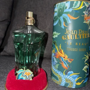 Jean Paul Gaultier Le Beau Parfym - Säljer en elegant parfymflaska från Jean Paul Gaultier, 'Le Beau', med en unik design som liknar en manlig torso. Flaskan är grön och kommer med en dekorativ förpackning med tropiskt mönster och texten 'Paradise Garden'. Perfekt för den som vill ha en doft med stil och karaktär.
