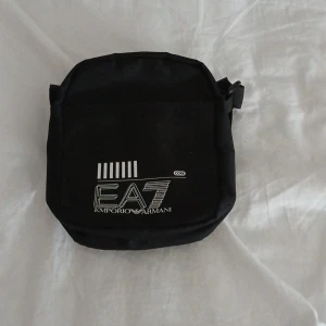 Svart axelväska från Emporio Armani - Snygg svart axelväska från Emporio Armani med logotypen EA7 på framsidan. Väskan har en justerbar axelrem och en praktisk dragkedja för säker förvaring. Perfekt för att bära med sig det nödvändigaste med stil.pris kan diskuteras