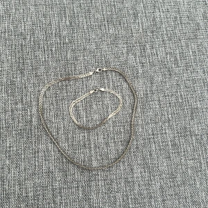 Silverfärgat halsband och armband - Elegant set med ett silverfärgat halsband och matchande armband. Båda smyckena har en enkel och stilren design med tunna kedjor och smidiga lås. Perfekt för att komplettera din look med en subtil glans.