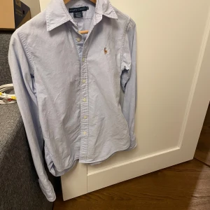 Ljusblå skjorta från Ralph Lauren - Säljer en stilren ljusblå skjorta från Ralph Lauren i 100% bomull. Skjortan har långa ärmar och klassisk krage med knappar framtill. Den ikoniska loggan är broderad på bröstet. Perfekt för en elegant look. Priset är 299 men inte hugget i sten!🫵Fråga om minsta lilla till exempel fler bilder✅Skjortan är i Ralph laurens storlek 4 vilket motsvarar xs/s