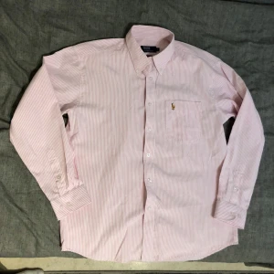 Rosa randig skjorta från Polo Ralph Lauren - Säljer en stilren rosa och vit randig skjorta från Polo Ralph Lauren. Skjortan är i 100% bomull och har långa ärmar med knappar vid ärmsluten. Den har en klassisk krage och en broderad logotyp på bröstet. Perfekt för en snygg och avslappnad look.