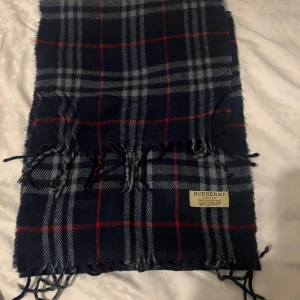 Rutig halsduk från Burberry - Snygg rutig halsduk från Burberry i klassiska färger. Den är tillverkad av 100% lammull och har fransar i ändarna. Perfekt för att hålla dig varm och stilren under kyliga dagar. Skriv vid eventuell frågor. Pres kan diskuteras 